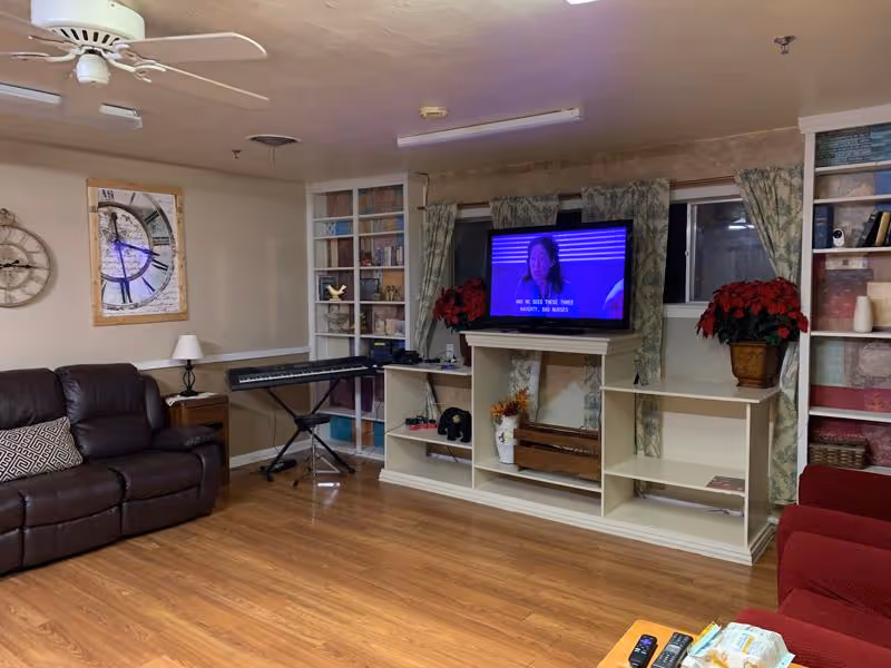 Communal living room with a TV on a white shelving unit, leather couch, keyboard, bookshelves, and wood flooring.