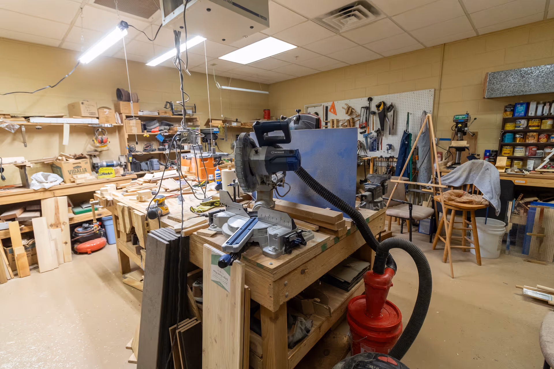 A woodworking workshop with various tools and materials. There is a large workbench in the center cluttered with wood pieces, a miter saw, and other woodworking tools. Shelves and pegboards on the walls hold additional tools, supplies, and containers. The room is well-lit with fluorescent lights on the ceiling.