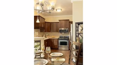 A modern kitchen with wooden cabinets, stainless steel appliances including a microwave and oven, and a dining table set with plates, glasses, and napkins. The room is well-lit with ceiling lights and pendant lighting.