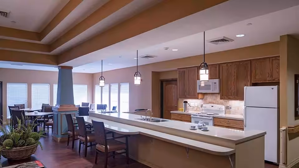 Interior view of a senior living facility kitchen and dining area with wooden cabinets, white appliances including a refrigerator, stove, and microwave, a long countertop with bar stools, pendant lights hanging from the ceiling, and a dining table with chairs near windows with blinds.