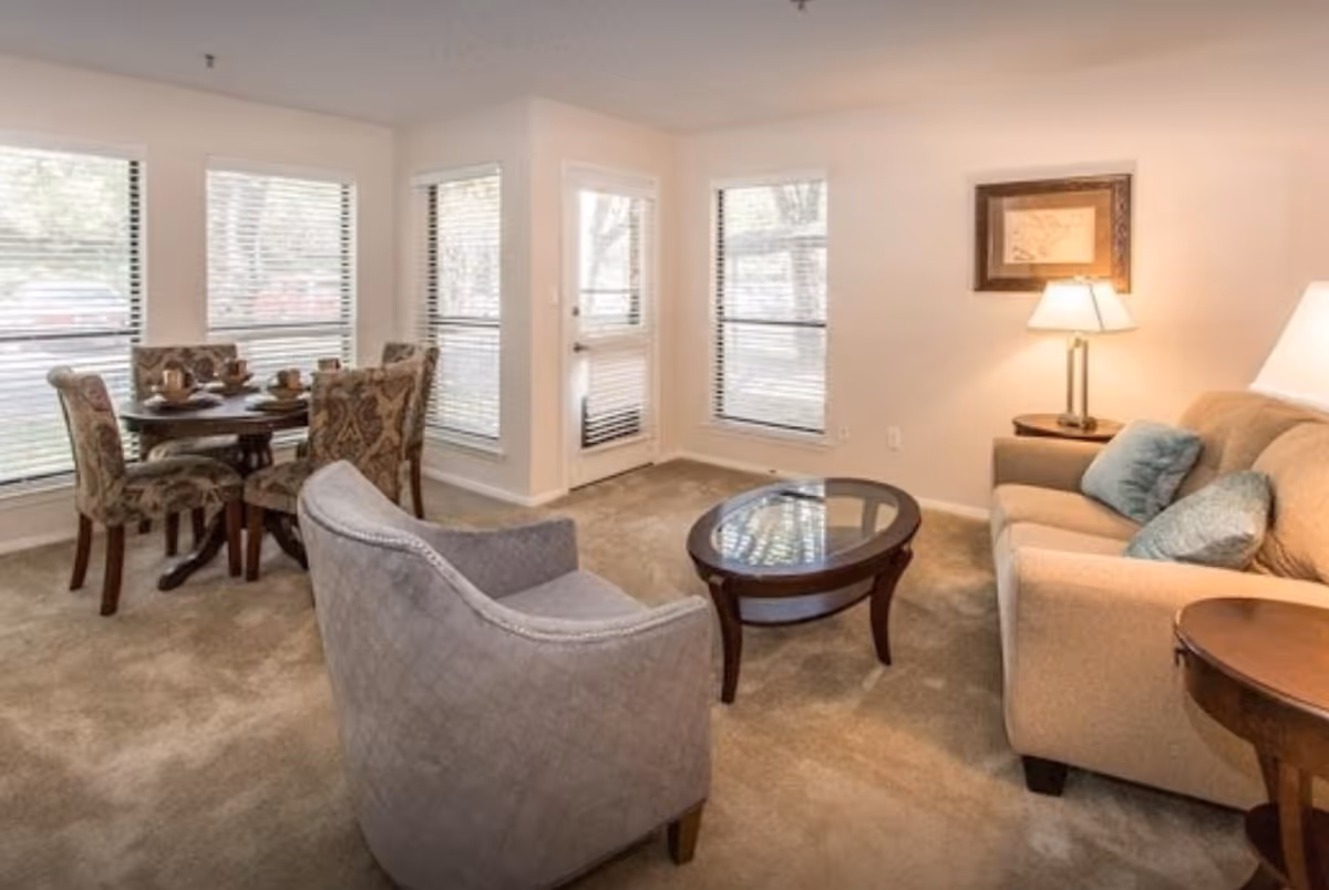 A cozy living room and dining area in a retirement community. The living room features a beige sofa with blue cushions, a glass-top coffee table, a side table with a lamp, and a gray upholstered armchair. The dining area has a round wooden table with four patterned chairs. Large windows with blinds let in natural light, and a door leads outside.