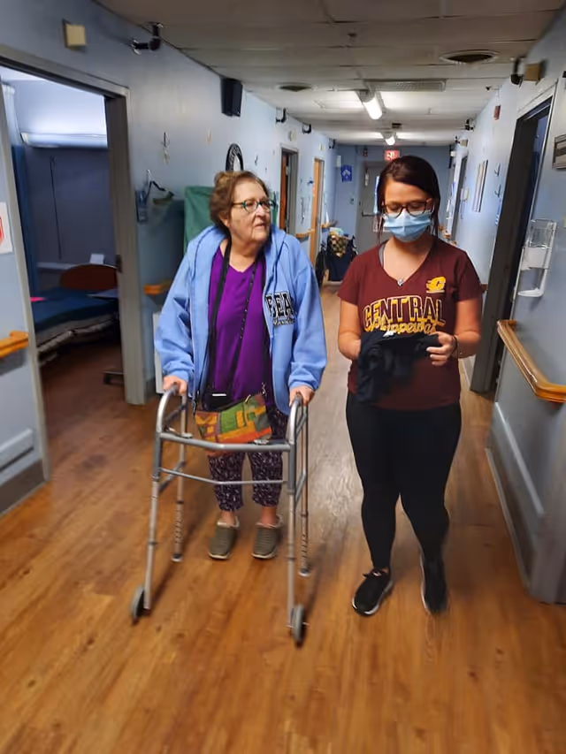 An elderly woman using a walker is walking down a hallway in a nursing facility accompanied by a younger woman wearing a mask and a Central Michigan University shirt. The hallway has wooden floors, handrails on the walls, and several doors leading to rooms.