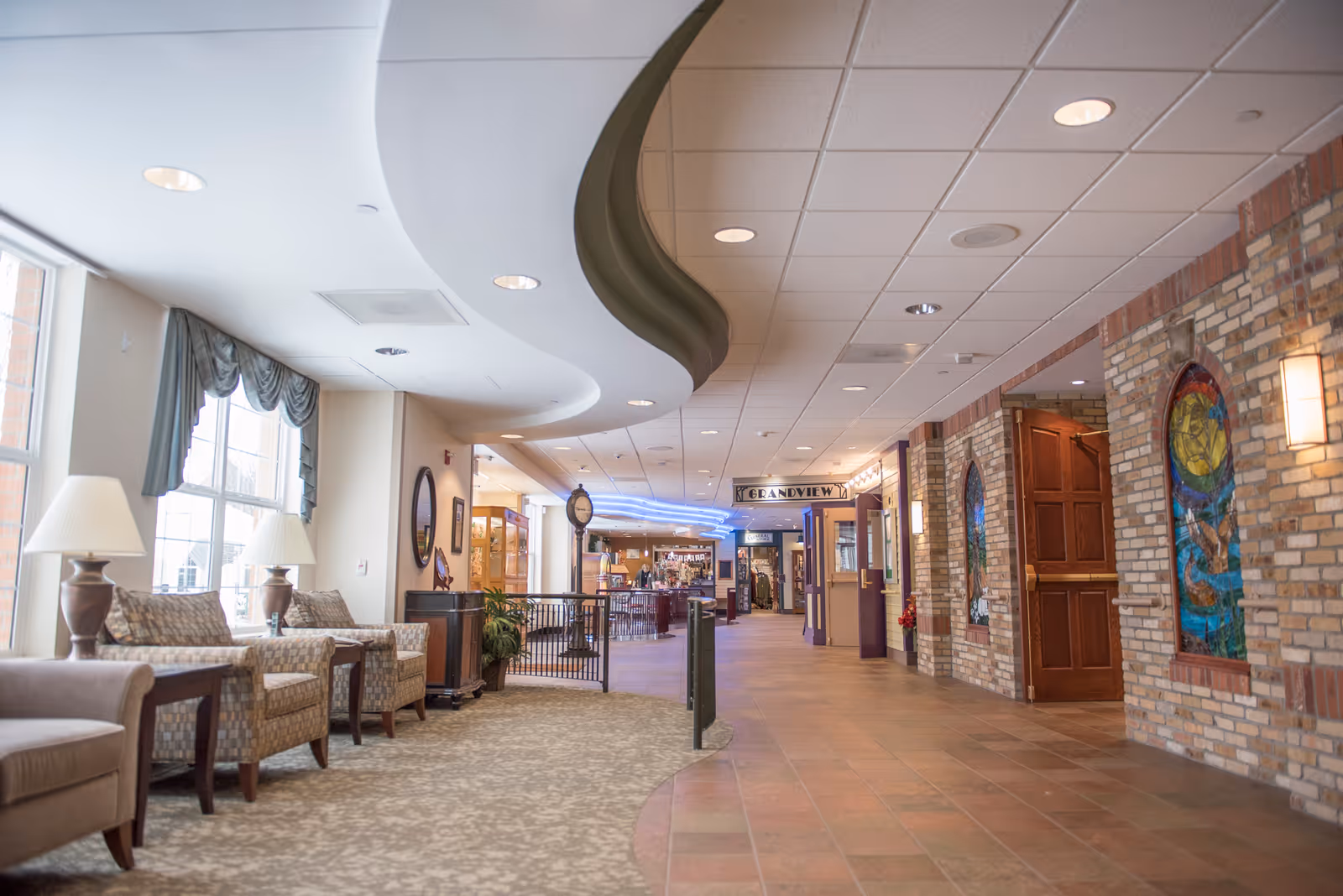 A spacious and well-lit hallway in a senior living facility with comfortable seating along the left side near large windows with curtains. The right side features a brick wall with colorful stained glass window decorations and a wooden door. The ceiling has recessed lighting and a curved design element. At the end of the hallway, there is a sign that reads 'GRANDVIEW' above a doorway.