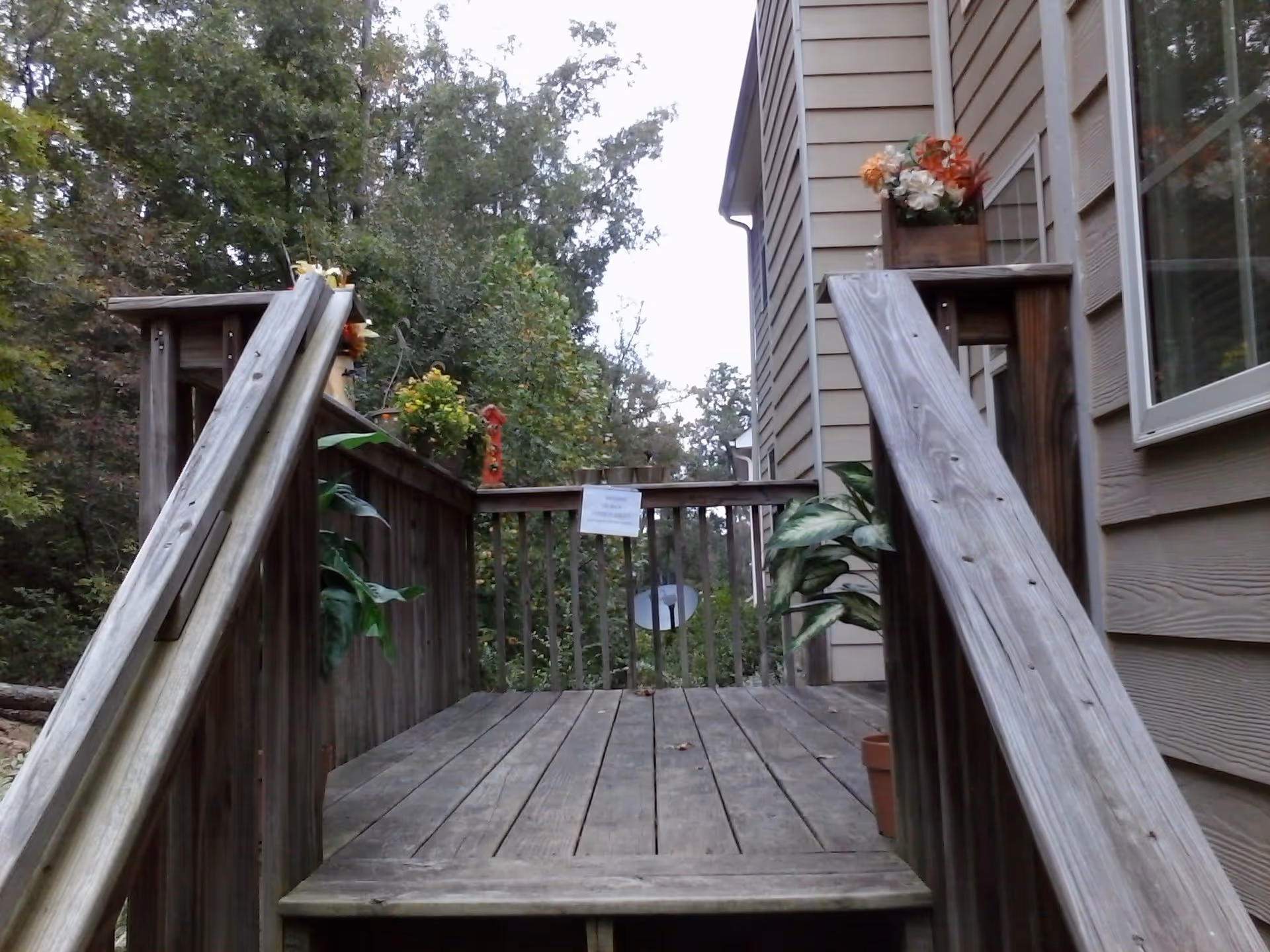 Wooden deck with railings and stairs leading up to a house exterior. The deck has potted plants and flower arrangements on the railings. Trees and greenery are visible in the background.
