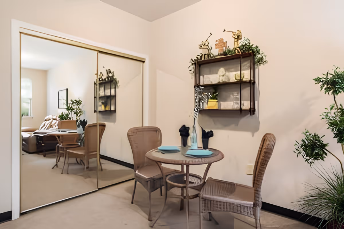 A small dining area with a round glass table set for two with blue plates and black napkins. Two wicker chairs are placed around the table. A wall-mounted shelf with decorative items and plants is above the table. A large mirrored sliding closet door reflects part of the room, showing a bed and additional furniture. There are green plants on the right side of the image, and the walls are painted light beige.