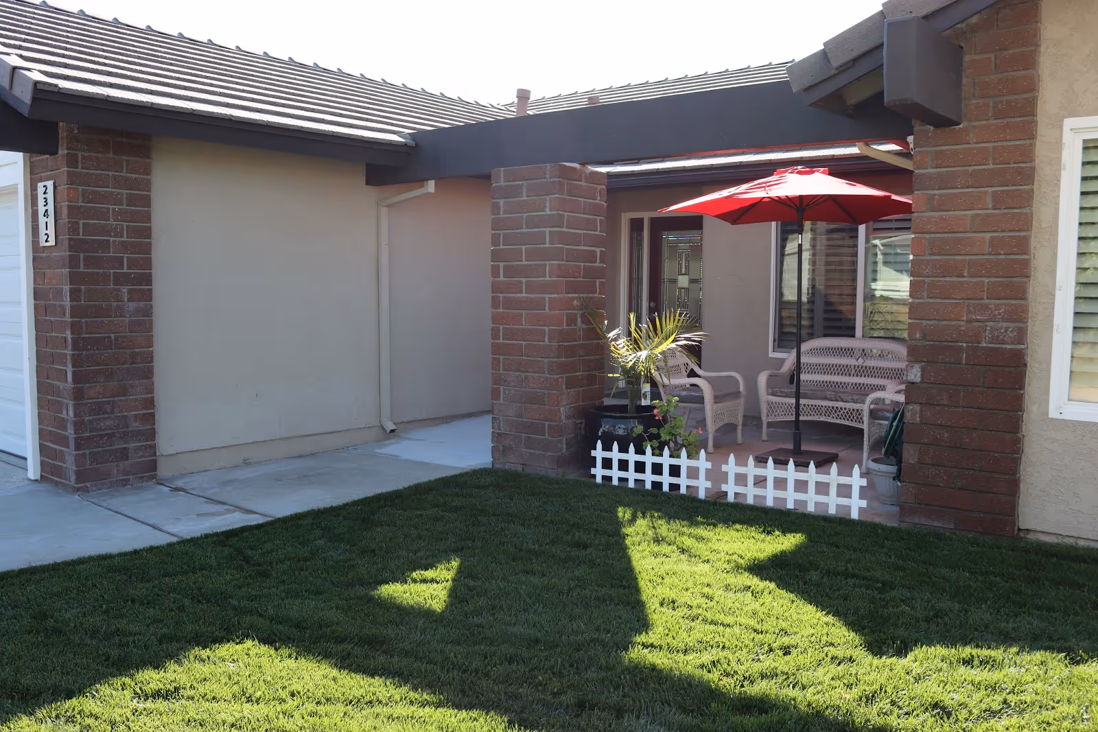 Small front patio of a single-story building with brick columns, a red umbrella over wicker chairs, potted plants, and a green lawn.