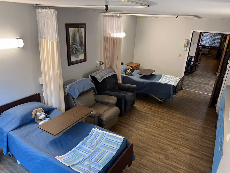 A shared bedroom in a senior living facility with two beds covered in blue blankets, each with a striped towel at the foot and a stuffed animal on top. Between the beds are two recliner chairs with a small wooden table in front of each. Privacy curtains hang from ceiling tracks between the beds. The room has wood flooring and neutral-colored walls with a framed picture on one wall. A hallway is visible in the background.