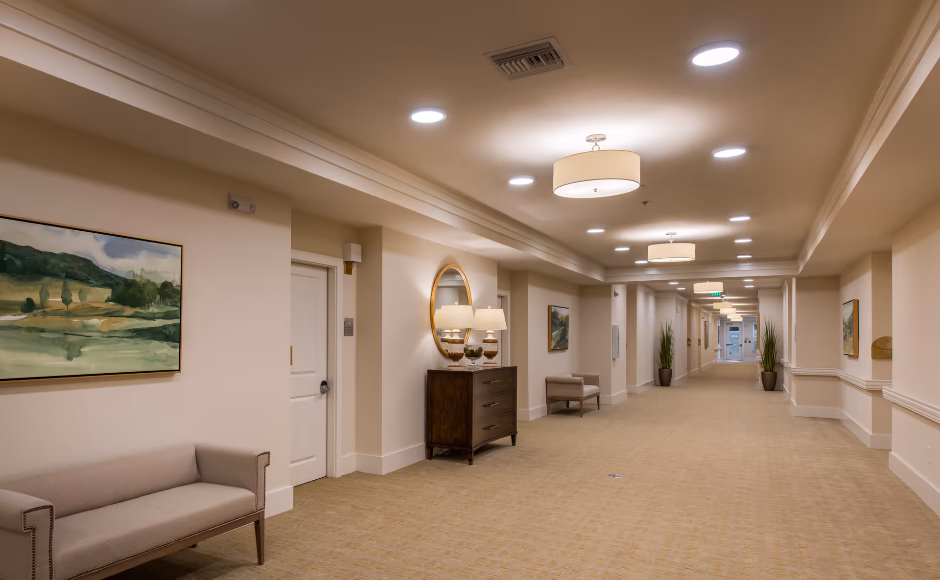 A long, well-lit hallway in a senior living facility with beige walls and carpet. The hallway features several pieces of furniture including a small sofa, a chest of drawers with a round mirror and lamps on top, and a single armchair. There are framed landscape paintings on the walls and potted plants near the far end of the corridor. Ceiling lights are evenly spaced along the hallway.