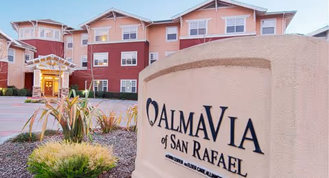 Exterior view of AlmaVia of San Rafael senior living facility showing a multi-story building with a landscaped area and a large sign with the facility's name in the foreground.