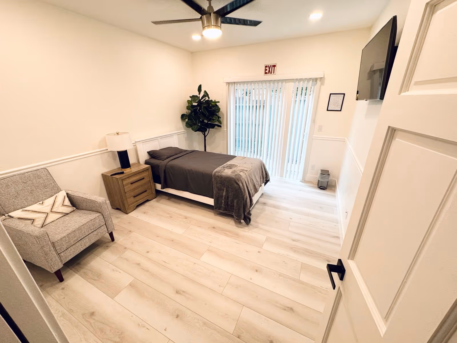 A clean and modern bedroom with light wood flooring, a single bed with dark gray bedding, a wooden nightstand with a lamp, a gray armchair with a decorative pillow, a ceiling fan with lights, a large plant in the corner, a wall-mounted TV, and sliding glass doors with vertical blinds leading outside. An exit sign is above the door.