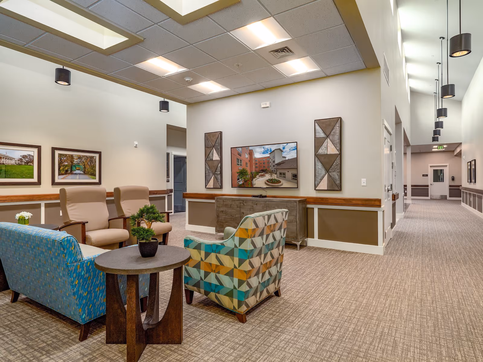 A bright and spacious senior living community lounge area with comfortable seating including two beige armchairs, a blue patterned sofa, and a multicolored geometric patterned armchair around a small round wooden table with a small potted plant. The walls are decorated with framed pictures and geometric wall art. The carpeted floor extends into a hallway with pendant lights hanging from the ceiling.