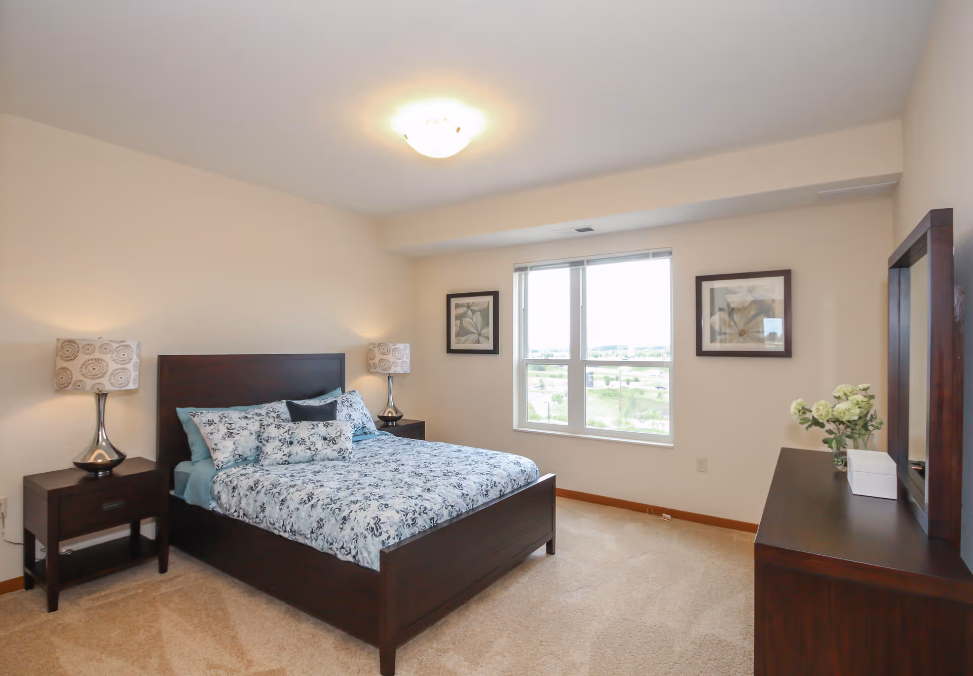 A bright and clean bedroom with a large window letting in natural light. The room features a dark wood bed frame with floral bedding, two matching nightstands with decorative lamps, a dresser with a mirror, and framed floral artwork on the walls. Beige carpet covers the floor.