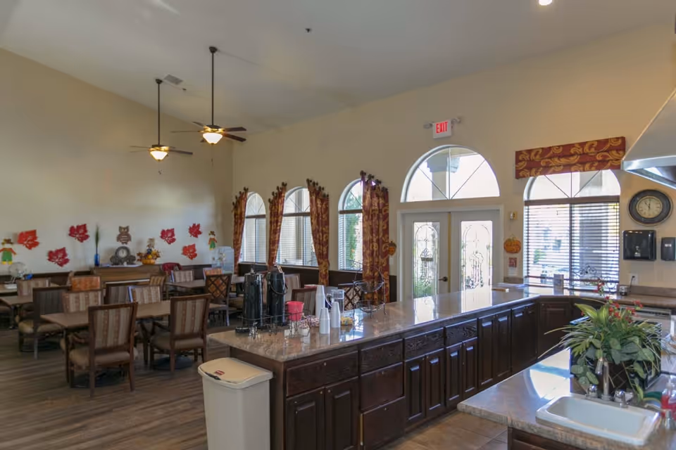 Spacious assisted living dining area with multiple tables and chairs, a long serving counter/island, arched windows and decorative curtains.