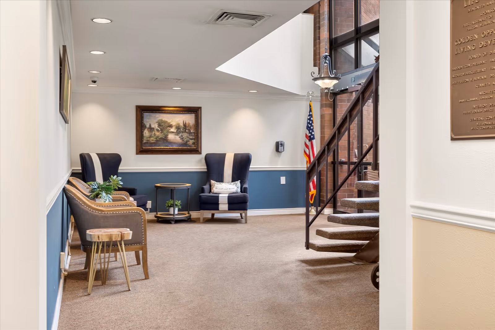 Bright seating area in a senior living facility with armchairs, small tables, a painting on the wall, and a spiral staircase by an American flag.