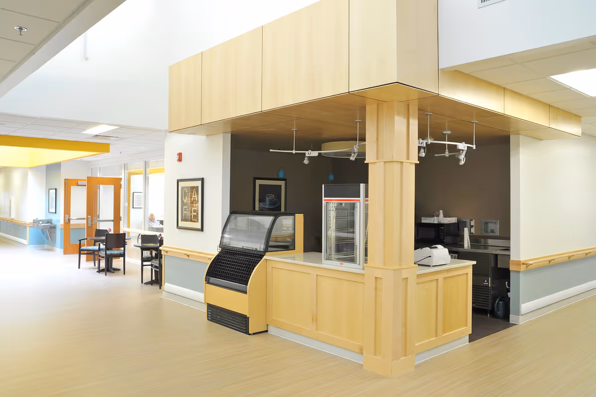 A small café or snack bar area inside a senior living facility with light wood cabinetry and counters, an empty display case, a cash register, and a hallway with tables and chairs visible in the background.