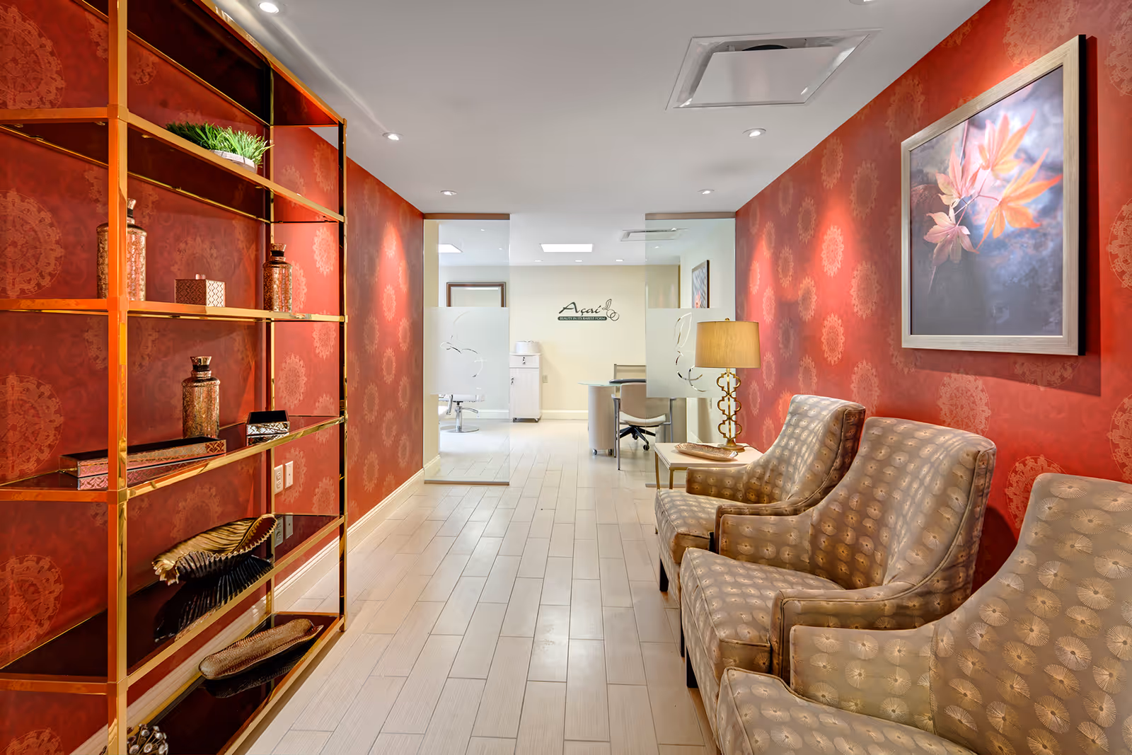 A decorated interior waiting area with patterned armchairs, a side table and lamp, gold shelving with decor, and red patterned wallpaper leading to an office corridor.