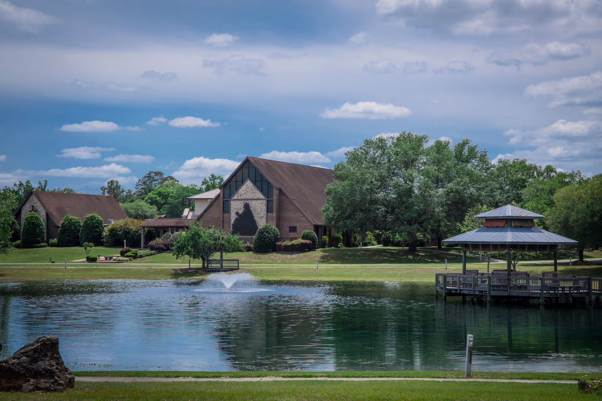 A peaceful outdoor scene at Advent Christian Village featuring a large pond with a water fountain in the center, a wooden gazebo on the right side over the water, and buildings with pitched roofs surrounded by trees and greenery under a partly cloudy sky.