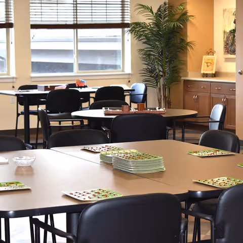 A dining room with multiple tables and black chairs arranged around them. Plates with a green and white pattern are set on the tables. There is a large window with blinds, a tall potted plant, and wooden cabinets along one wall.