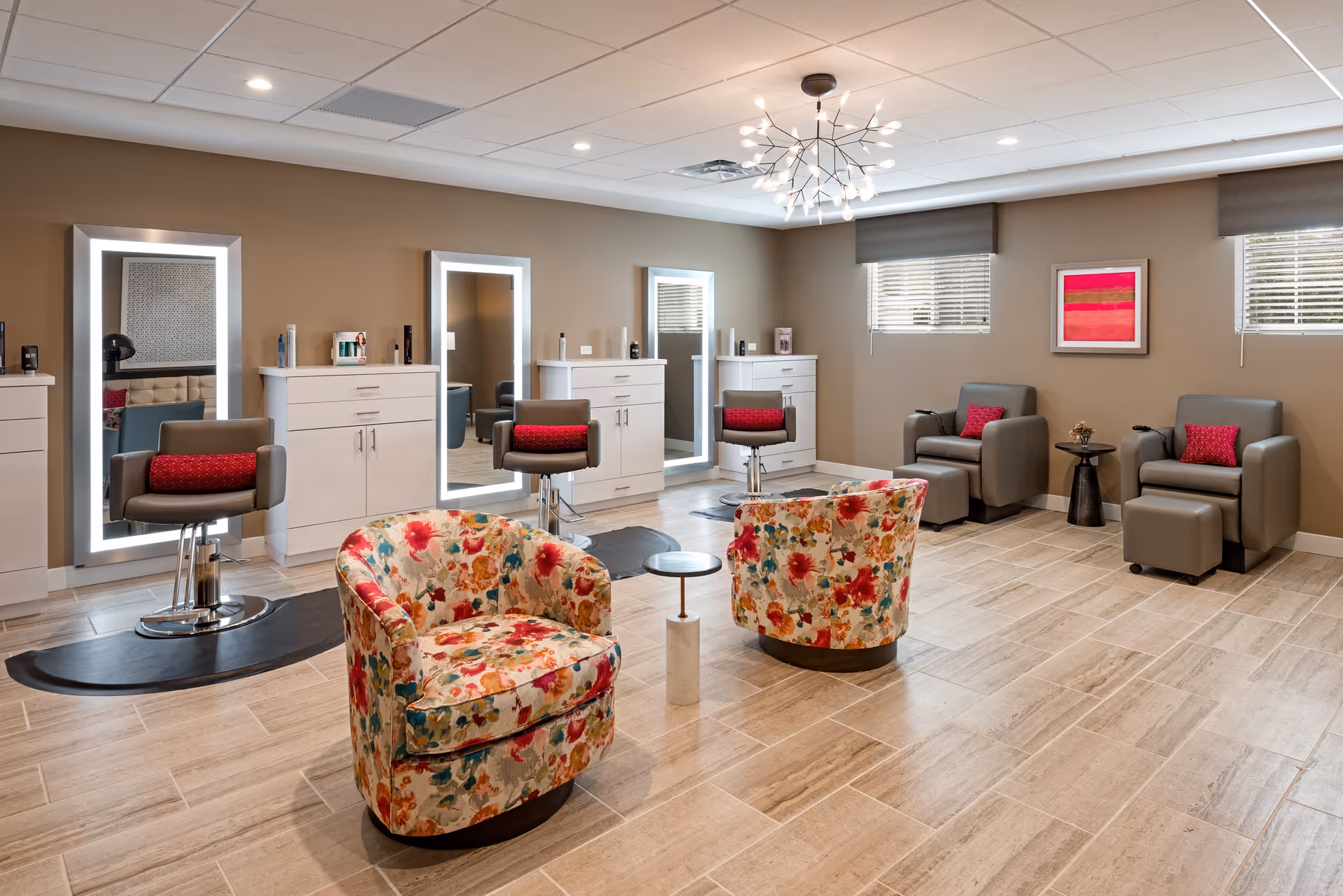 A modern salon area with three styling stations featuring large illuminated mirrors, white cabinetry, and brown salon chairs with red cushions. In the foreground, there are two floral-patterned armchairs with a small round table between them. To the right, there are two gray armchairs with matching ottomans and red cushions, positioned near two windows with blinds and a colorful abstract painting on the wall. The room has beige walls, light wood flooring, and a decorative ceiling light fixture.