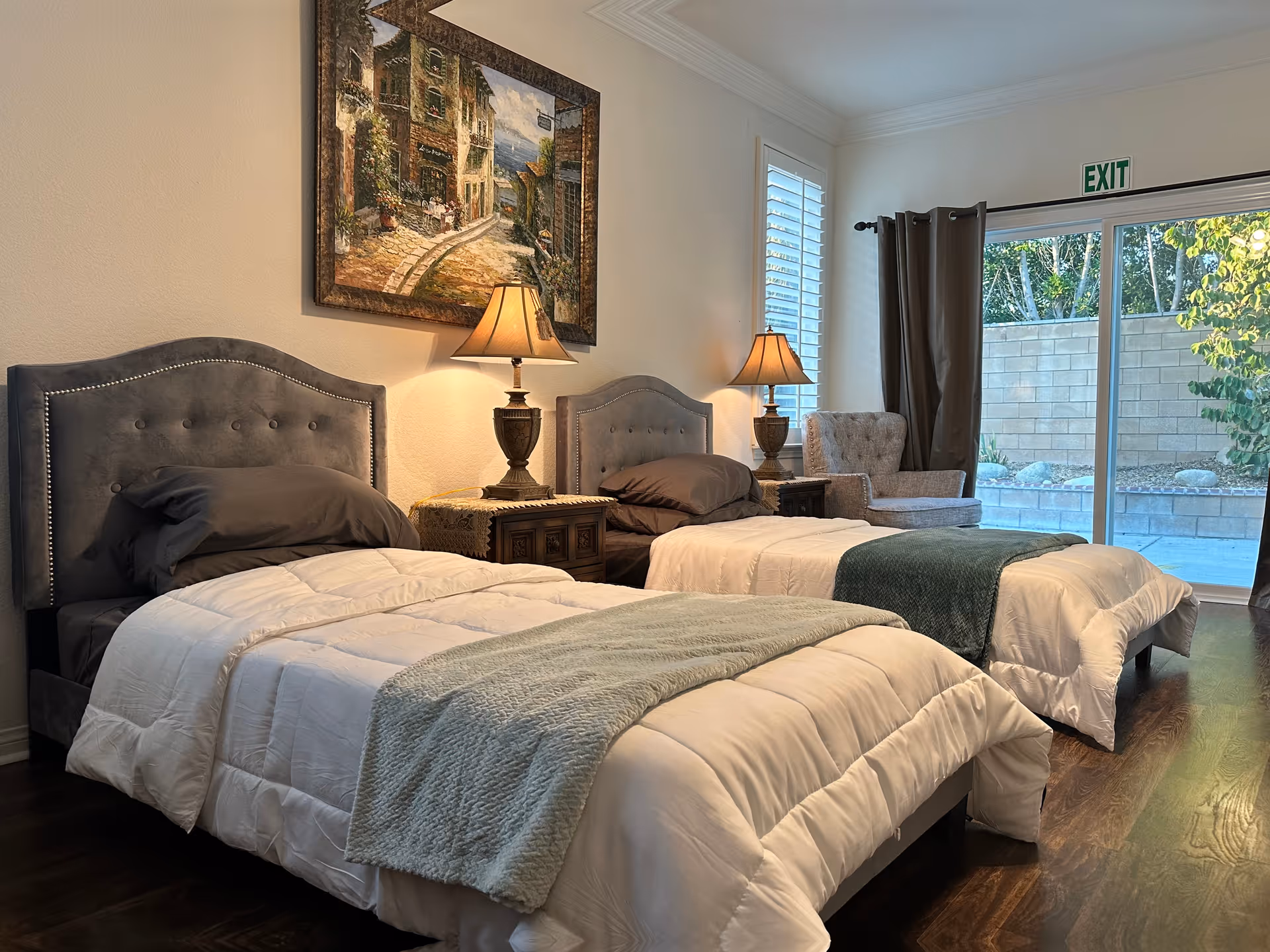 A cozy bedroom with two twin beds featuring gray upholstered headboards, white comforters, and dark gray pillows. Between the beds is a wooden nightstand with a decorative lamp. A framed painting hangs on the wall above the nightstand. There is a cushioned armchair near a window with white shutters and a sliding glass door leading outside, with an exit sign above it.