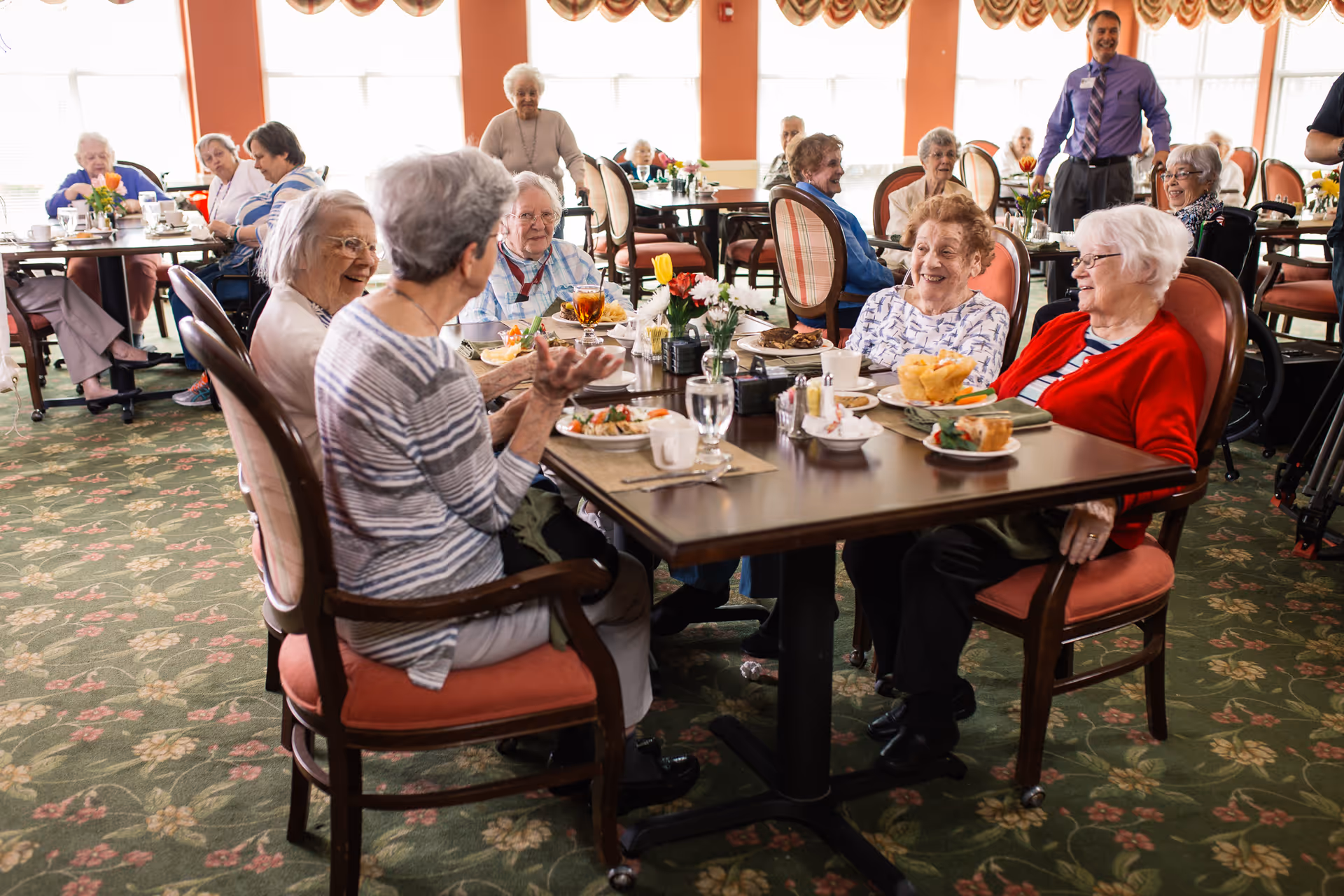 A group of elderly people sitting around tables in a dining room, enjoying a meal and engaging in conversation. The room has large windows with curtains, floral carpet, and several tables with chairs. A staff member in a purple shirt is standing in the background smiling.