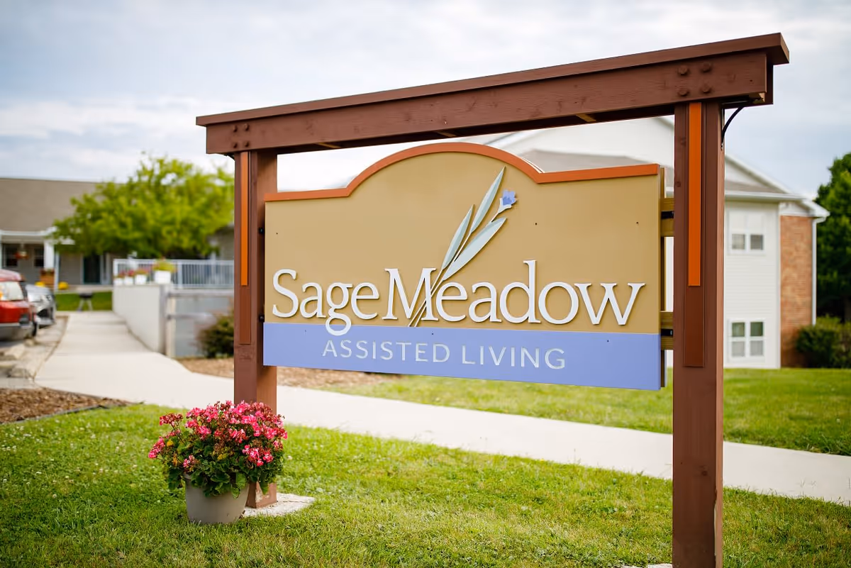 A large outdoor sign for Sage Meadow Assisted Living mounted on a wooden frame with a pot of pink flowers at its base. In the background, there is a sidewalk, some parked cars, and parts of the assisted living facility buildings with green grass and trees.