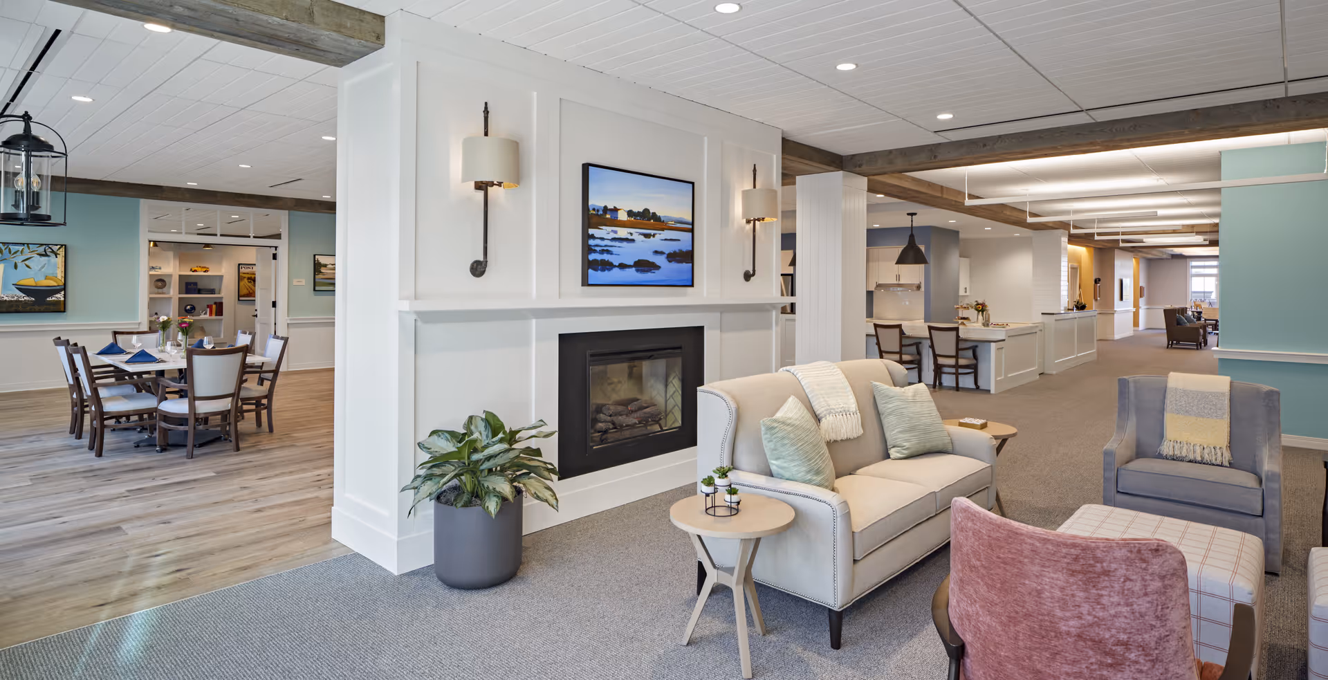 A spacious and well-lit senior living facility common area featuring a cozy seating arrangement with a beige sofa, armchairs, and a small round table. A modern fireplace is built into a white partition wall with a landscape painting above it. In the background, there is a dining area with a wooden table and chairs, and additional seating and decor throughout the open interior space.