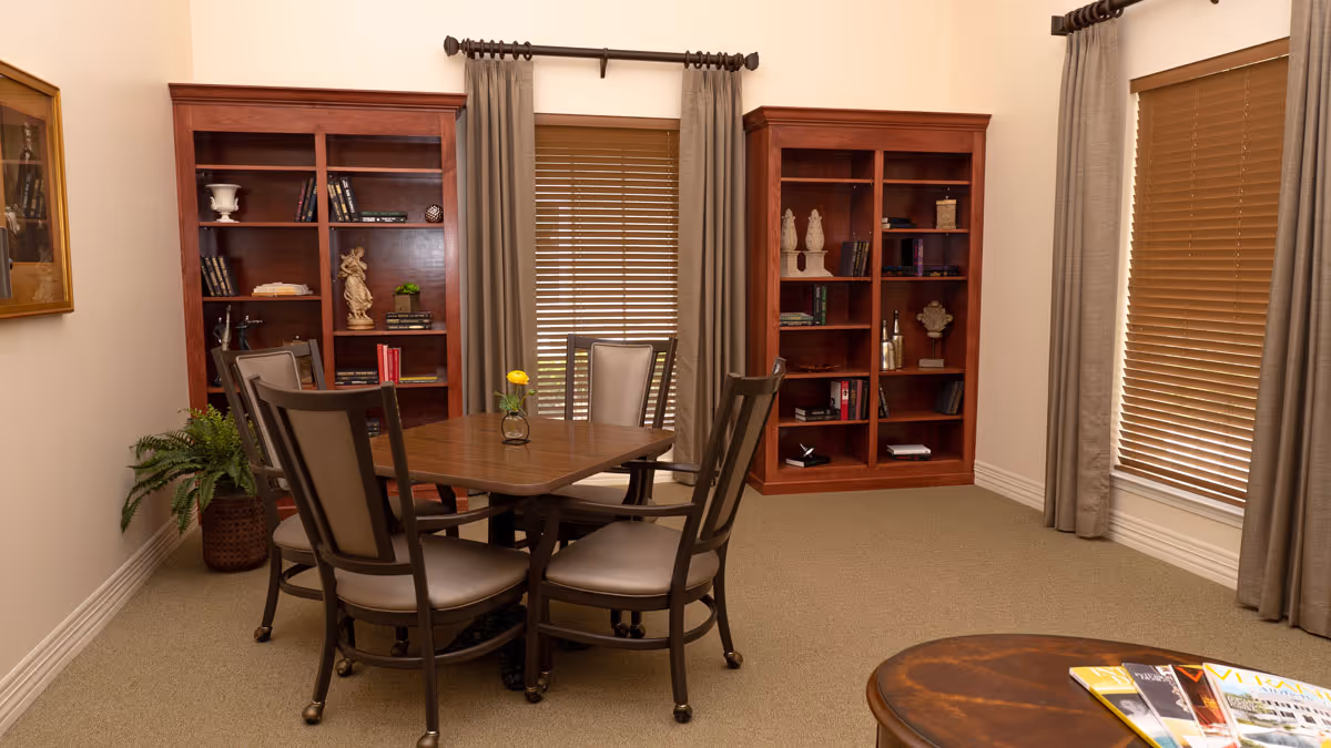 A cozy room with a square wooden table surrounded by four cushioned chairs. Behind the table are two wooden bookshelves filled with books and decorative items. The room has beige walls, carpeted floor, and two windows with brown blinds and gray curtains. A small plant is placed in the corner near one bookshelf, and a round wooden coffee table with magazines is partially visible in the foreground.