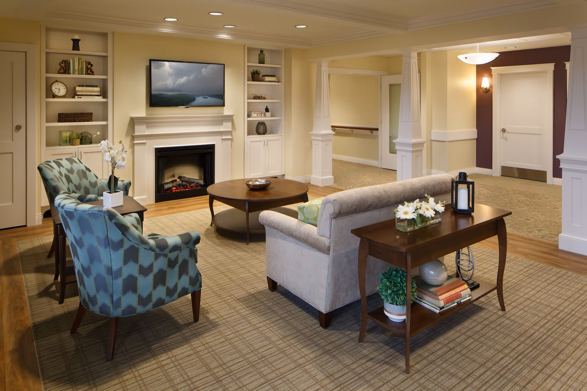 A cozy living room area with a beige sofa, two patterned armchairs, a round wooden coffee table, and a wooden side table with decorative items including flowers and books. The room features built-in white shelves flanking a fireplace with a TV mounted above it. The walls are painted in warm tones with recessed lighting in the ceiling.