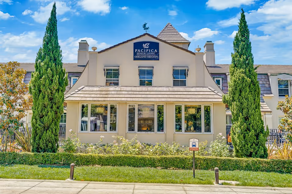Front exterior view of a senior living facility named Pacifica Senior Living Oakland Heights, featuring a beige building with multiple windows, two tall green trees on either side, and a well-maintained garden with bushes and flowers in front.