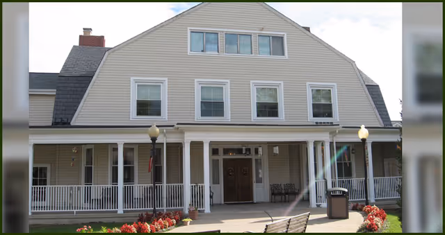 Front exterior of a multi-story assisted living building with a covered porch, white columns, flower beds, a bench, and double entry doors.