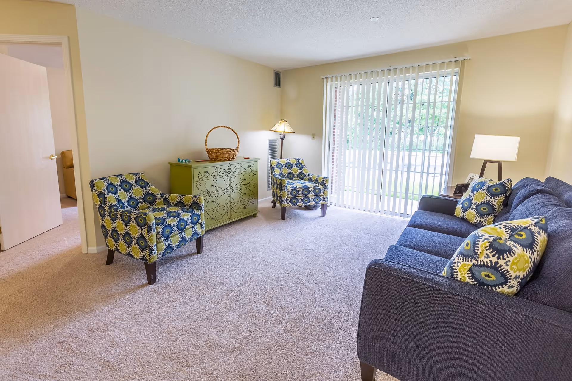 A bright living room with beige walls and carpet, featuring a blue sofa with patterned cushions, two matching patterned armchairs, a green decorative cabinet with a basket on top, a floor lamp, and a table lamp. Large vertical blinds cover a sliding glass door that leads outside.