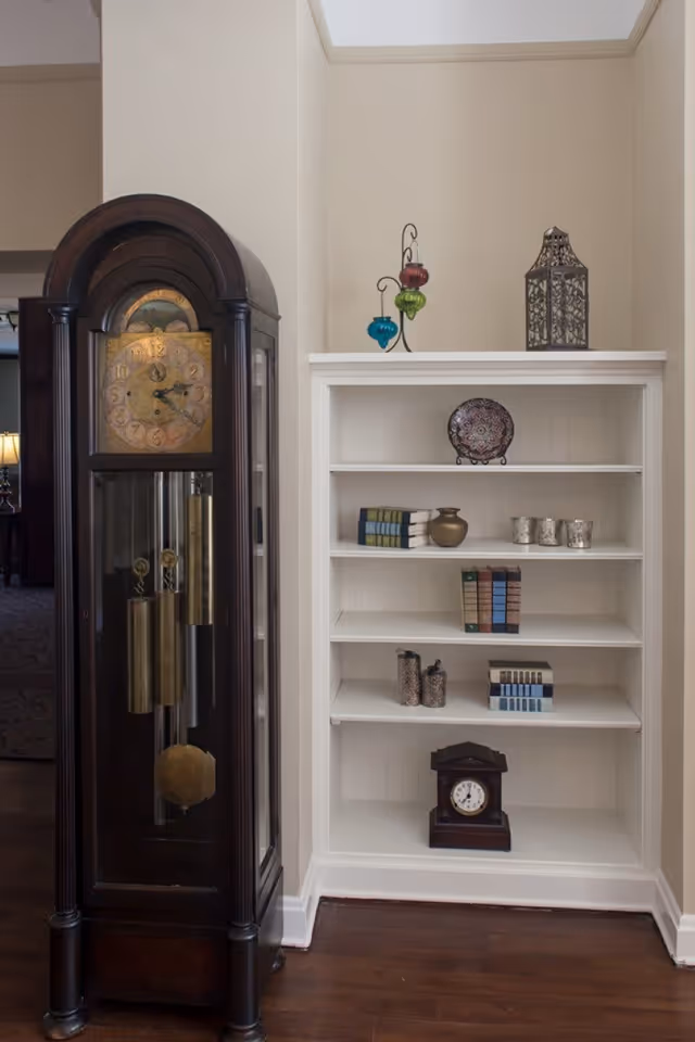 A corner of a room featuring a tall, dark wooden grandfather clock with brass weights and pendulum on the left, and a white built-in bookshelf on the right. The bookshelf contains decorative items including books, a small clock, a decorative plate, a vase, candle holders, and colorful hanging glass ornaments on top. The room has light-colored walls and dark wooden flooring.
