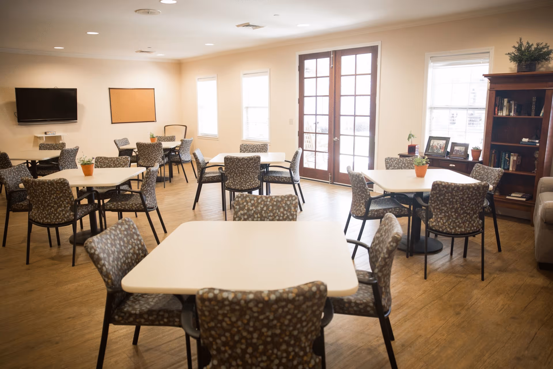 A bright and spacious common area with multiple square tables surrounded by patterned chairs. The room features wooden flooring, large windows, and French doors letting in natural light. There is a wall-mounted TV, a cork bulletin board, and a wooden bookshelf with books and framed photos. Small potted plants are placed on the tables and window sills.