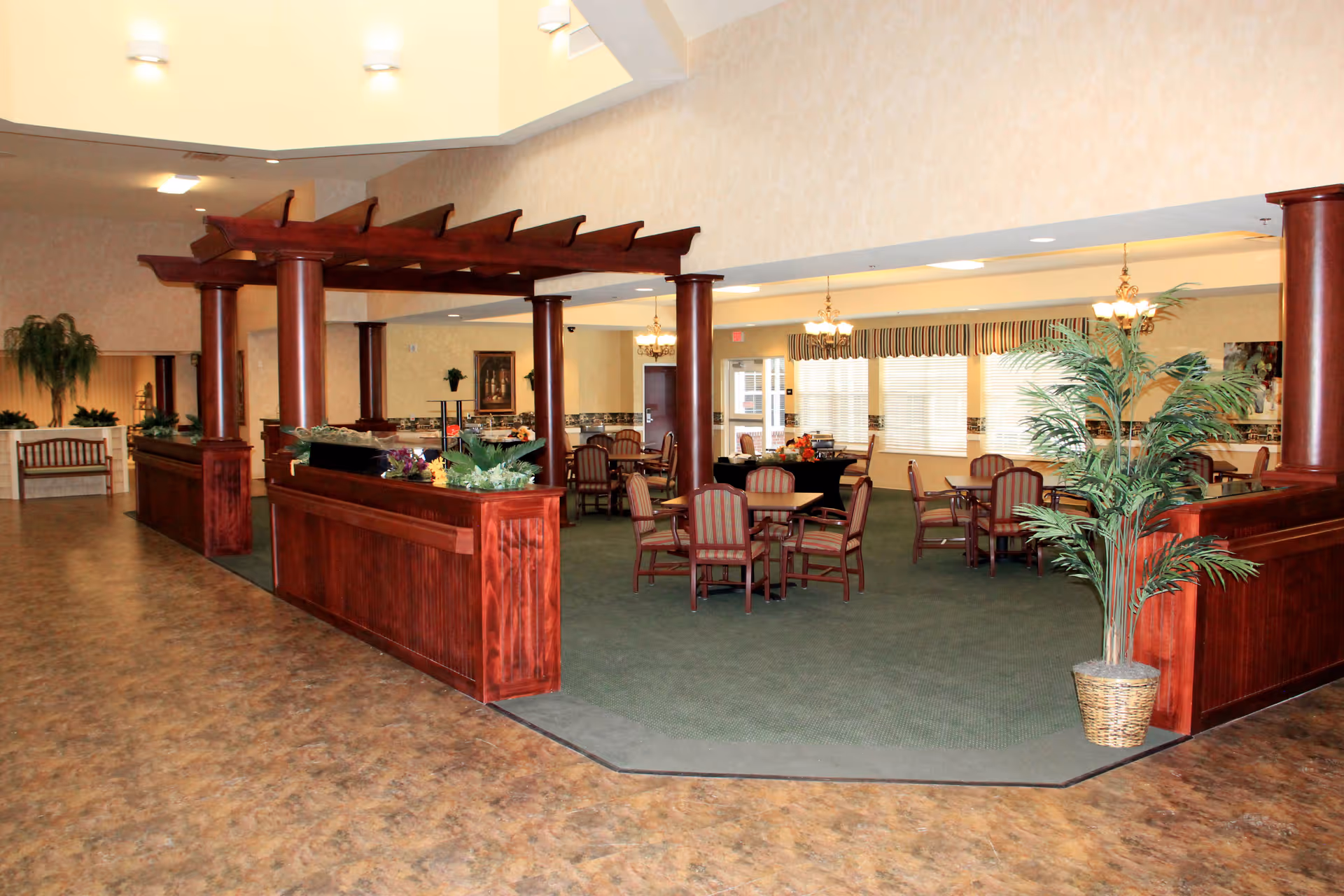 Interior view of a senior living facility dining area with wooden tables and chairs arranged on a green carpet. The space features wooden columns and a pergola-like structure with plants on the ledges. There are large windows with striped valances, chandeliers hanging from the ceiling, and a potted plant in the foreground.