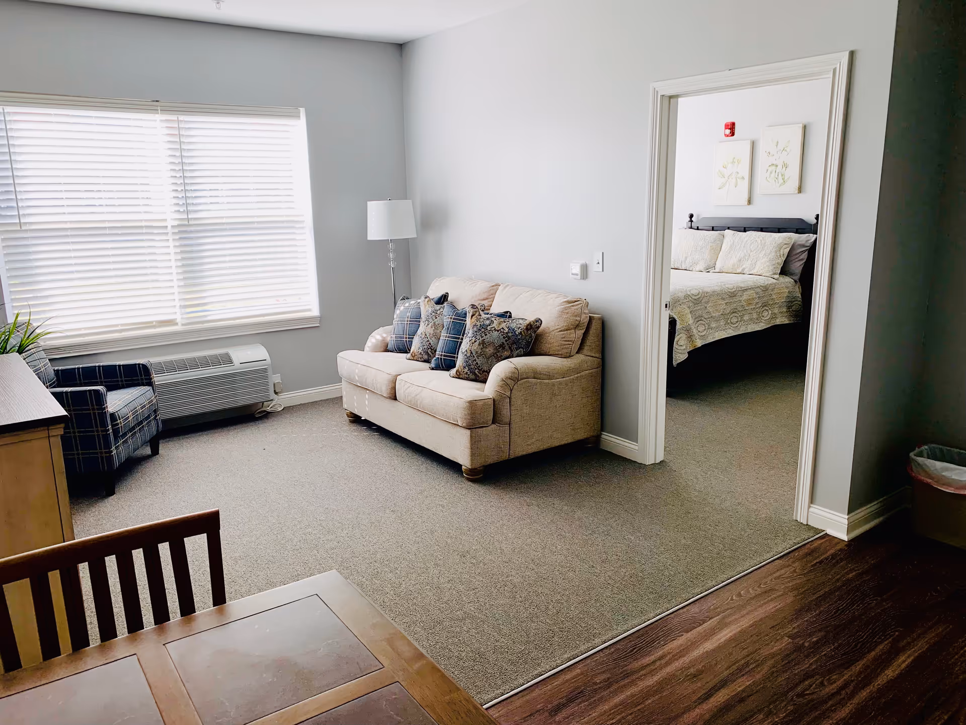 A bright living room with a beige sofa adorned with patterned pillows, a plaid armchair, a floor lamp, and a window with blinds. Adjacent to the living room is a bedroom with a bed covered in a light-colored quilt and two framed botanical prints on the wall.