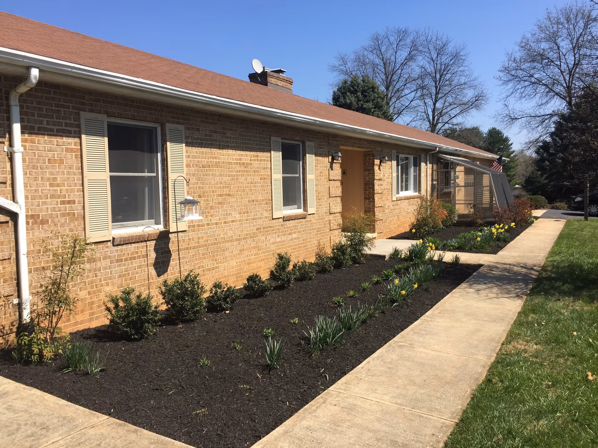 Single-story brick building with a concrete walkway, landscaped flower beds and a front entrance under a blue sky.