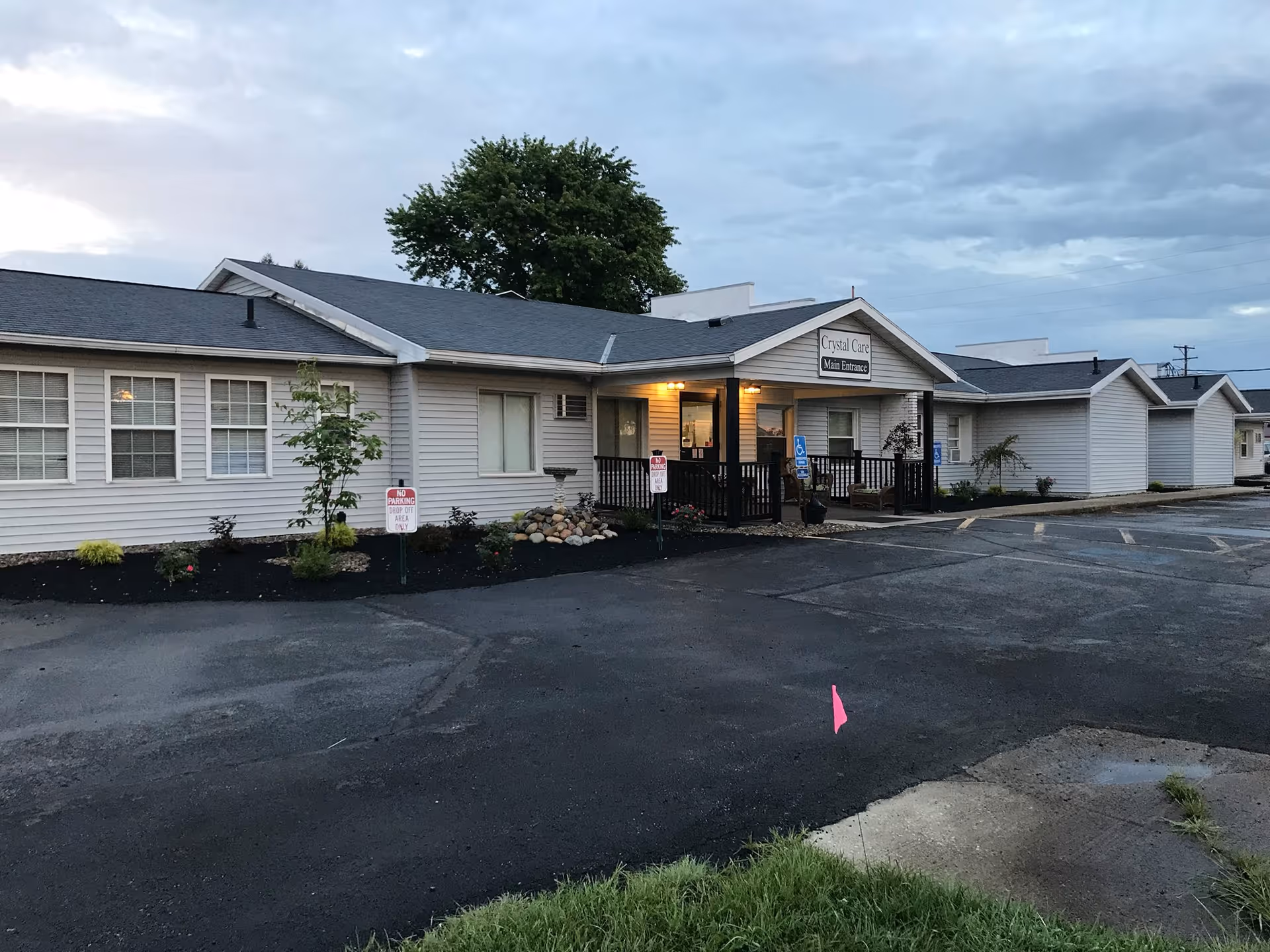 Exterior view of a single-story building with beige siding and a dark roof, featuring a small porch with lights on and a sign above the entrance that reads 'Crystal Care Main Entrance'. There are small landscaped areas with plants and a parking lot in front, including handicap parking spaces.