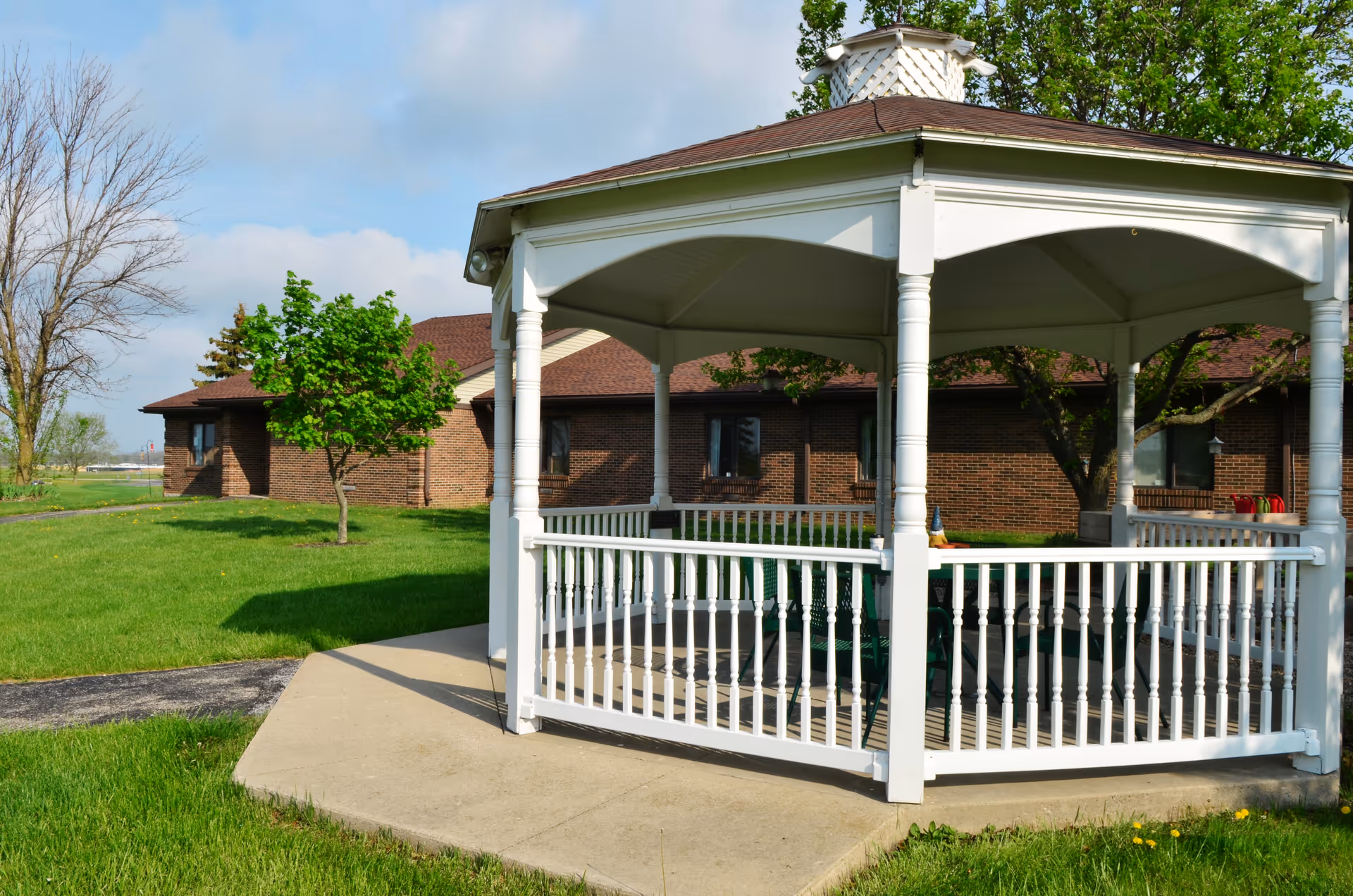 A white gazebo on a concrete pad sits on a green lawn in front of a single-story brick building with trees and a walking path.