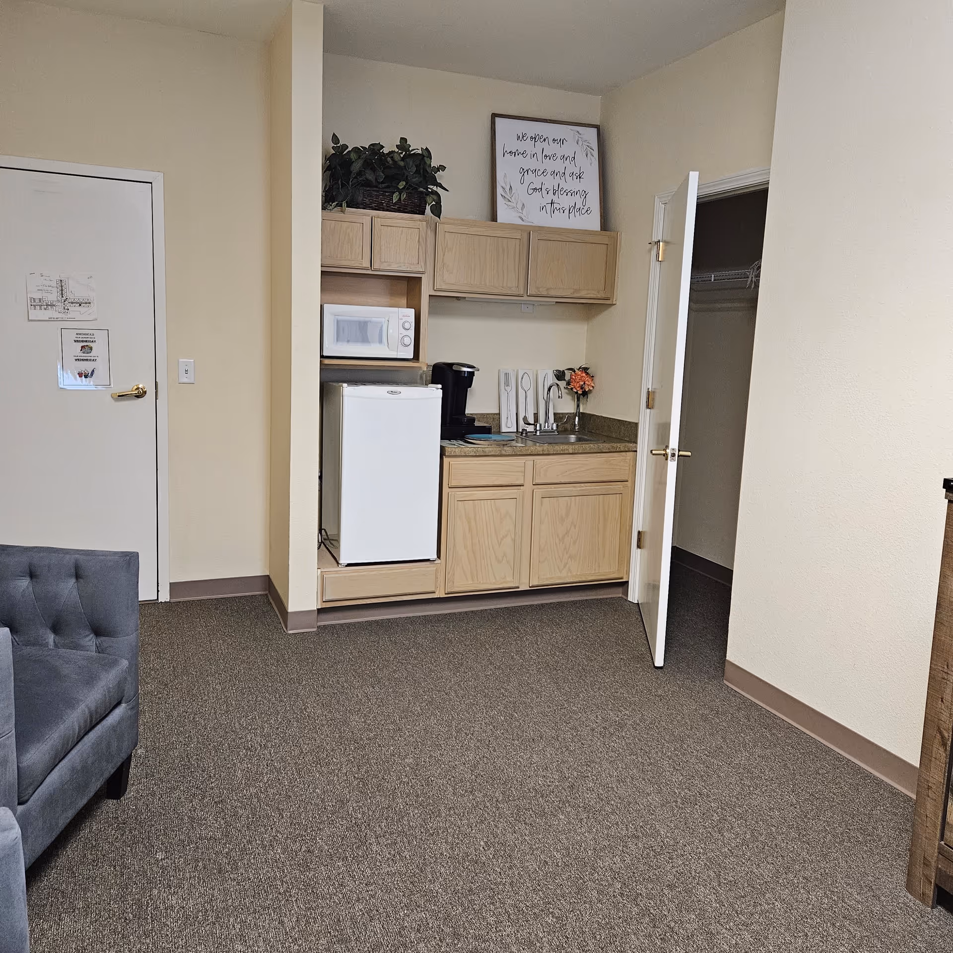 A small kitchenette area in a senior living facility room with light wood cabinets, a white mini refrigerator, a microwave, a coffee maker, and a small sink. There is a gray upholstered chair partially visible on the left and an open door leading to a closet on the right. A framed sign with a quote and a potted plant are placed on top of the cabinets.