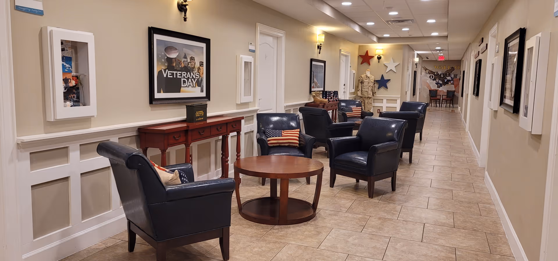 A hallway in a senior living facility with several black leather armchairs arranged around round wooden tables. The walls are decorated with framed pictures, including one with the text 'Veterans Day,' and patriotic-themed pillows are placed on the chairs. The floor is tiled, and the hallway is well-lit with wall sconces and ceiling lights.