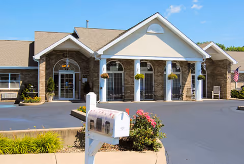 Front exterior view of a single-story senior living facility building with a covered entrance supported by white columns, brick walls, and large windows. There is a mailbox with a picture of the building on it, some landscaping with flowers and bushes, and an American flag on the right side.