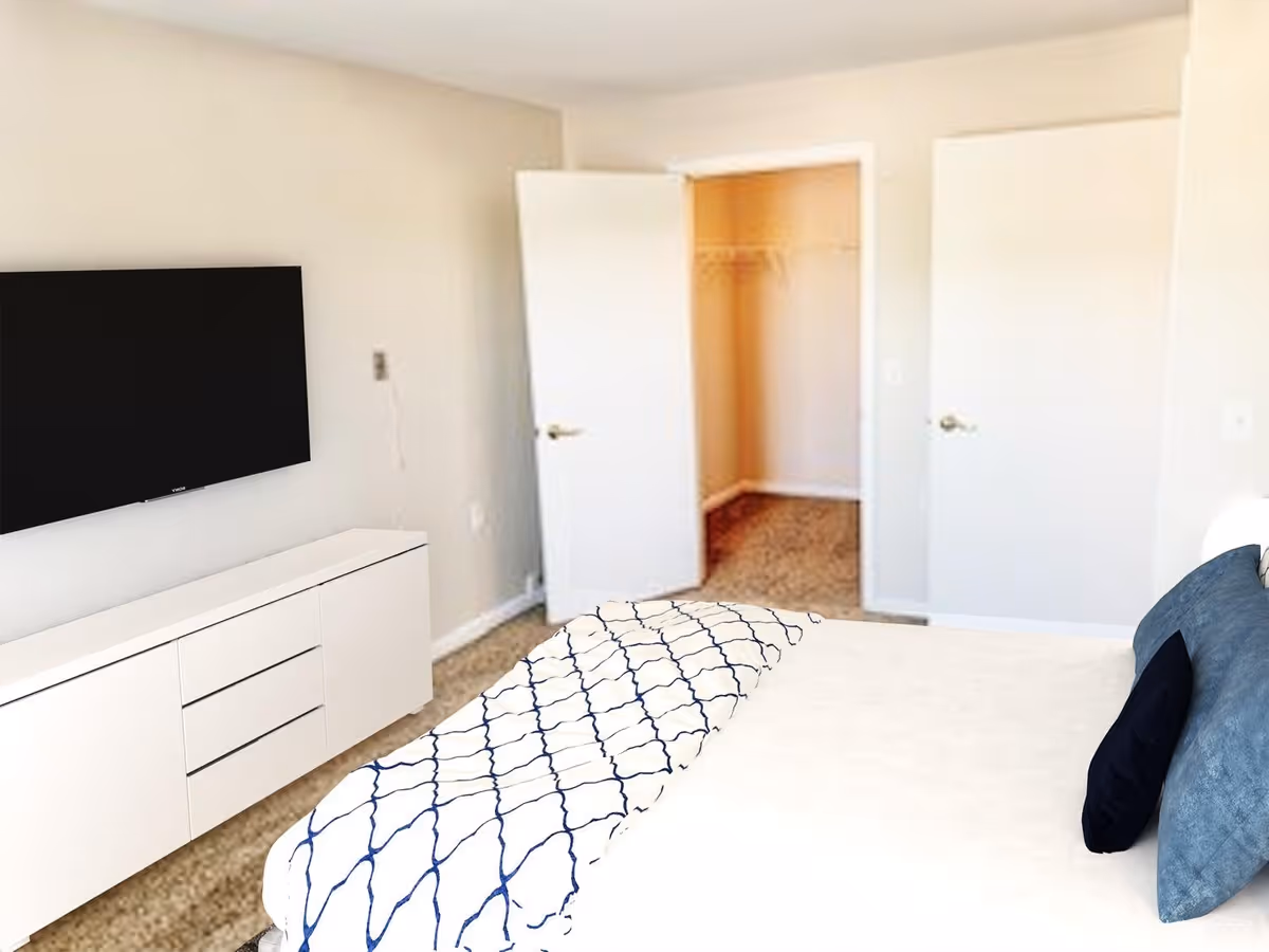 A bedroom with a bed featuring white and blue bedding and pillows, a white dresser with drawers, a wall-mounted flat-screen TV, and two open doors leading to a closet and another room. The walls are light-colored and the floor is carpeted.