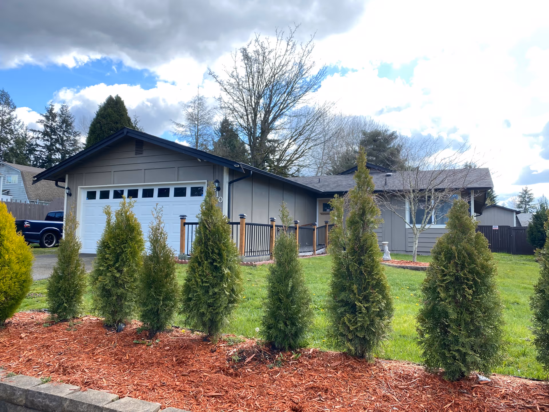 Single-story residential building with a gray exterior and a white garage door, surrounded by a green lawn and a row of small evergreen trees planted in red mulch in the foreground. The sky is partly cloudy with patches of blue visible.