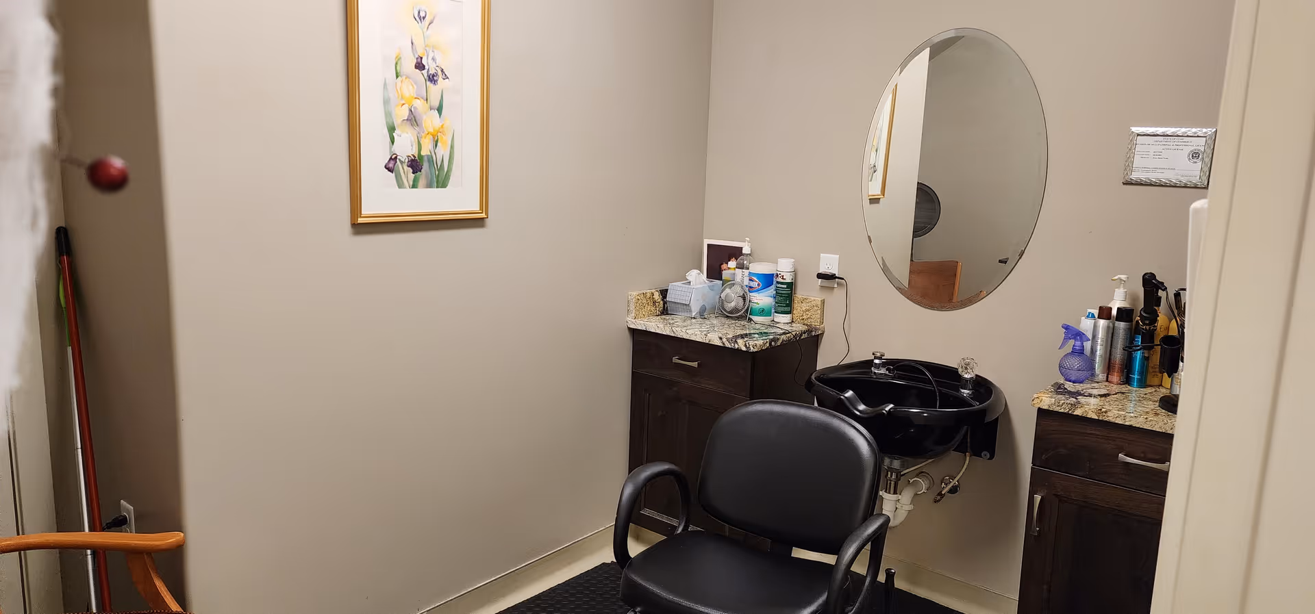 Interior of a small salon or grooming room with a black salon chair in front of a black sink. There are two dark wood cabinets with granite countertops on either side of the sink, holding various hair care products and supplies. A round mirror is mounted on the wall above the sink, and a framed floral picture hangs on the adjacent wall. A wooden chair and a broom are partially visible in the corner.