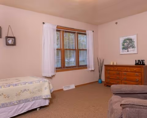 A cozy bedroom with a bed covered in a floral quilt on the left, a window with white curtains in the center, and a wooden dresser with candles and a decorative 'LIVE' sign on the right. A framed picture of a tree hangs above the dresser, and a comfortable armchair is partially visible in the foreground.