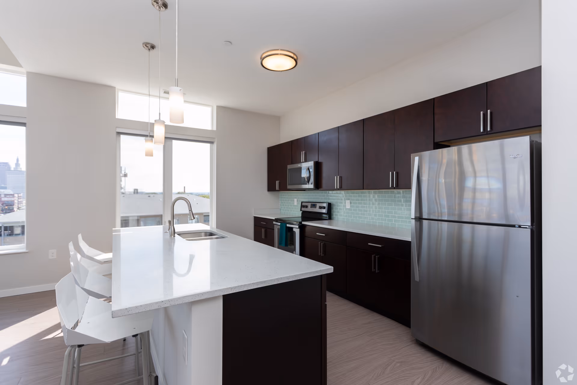 Bright modern kitchen with a white island and bar stools, dark cabinets, stainless appliances, and pendant lights.