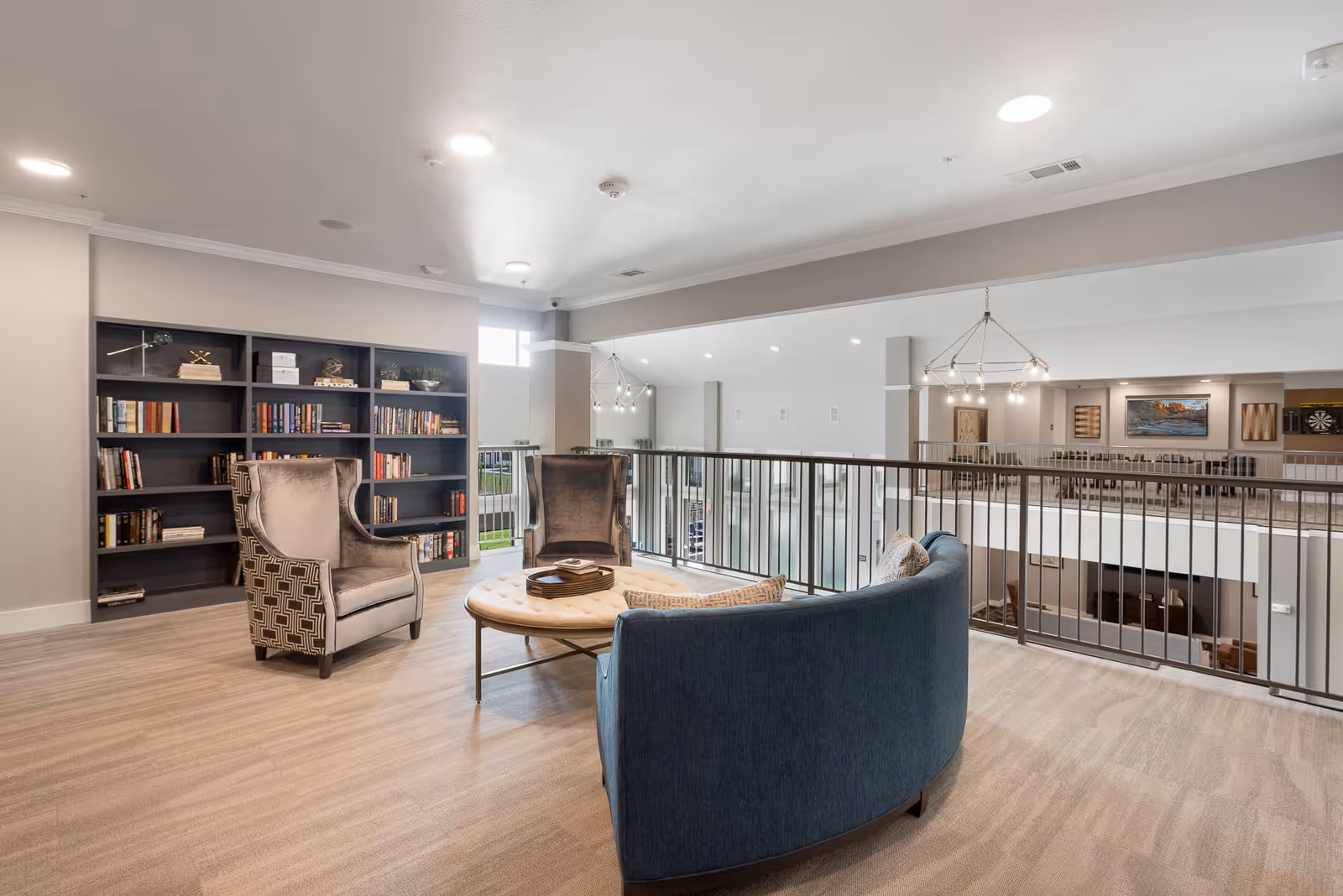 A bright and spacious lounge area with a curved blue sofa, two gray armchairs, and a round tufted ottoman coffee table. Behind the seating is a large bookshelf filled with books and decorative items. The area overlooks a lower level with railings and modern light fixtures hanging from the ceiling.