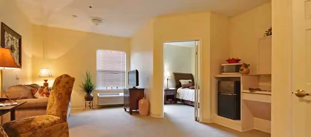 A cozy living area with a sofa, armchair, and table lamp on the left, a TV on a stand near a window with blinds, and an open doorway leading to a bedroom with a bed and nightstand. On the right, there is a small kitchenette area with a mini fridge and shelves.