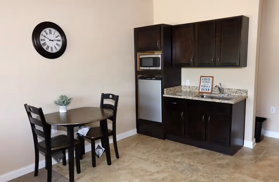Small kitchenette with dark wood cabinets, microwave, mini-fridge and sink beside a round table with three chairs and a wall clock.